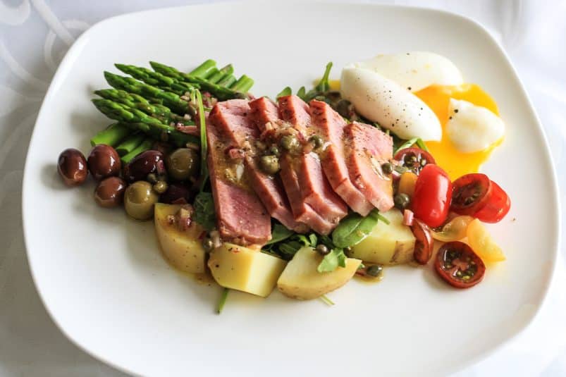 Nicoise salad composed of rare sliced tuna, nicoise olives, and runny egg arranged on a white plate.