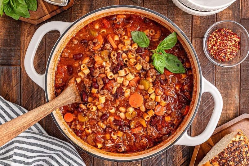 A steaming pot of savory pasta e fagioli soup (Olive Garden copycat) brimming with hearty vegetables, tender beans, and pasta is garnished with fragrant basil. Beside the wooden spoon rests a bowl of zesty red pepper flakes and warm bread, ready to complement this comforting dish.