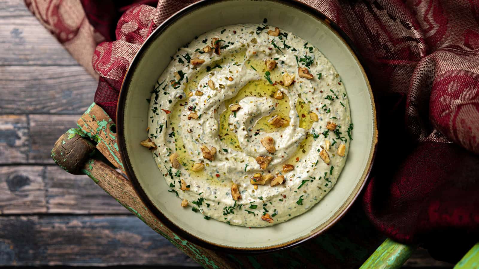 A bowl of whipped feta dip garnished with chopped herbs and nuts, drizzled with olive oil, set on a rustic wooden surface with a patterned cloth nearby.