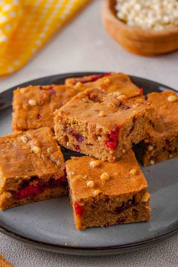 White chocolate raspberry blondies stacked on a plate. Some blurred white chocolate chips are visible in the background.