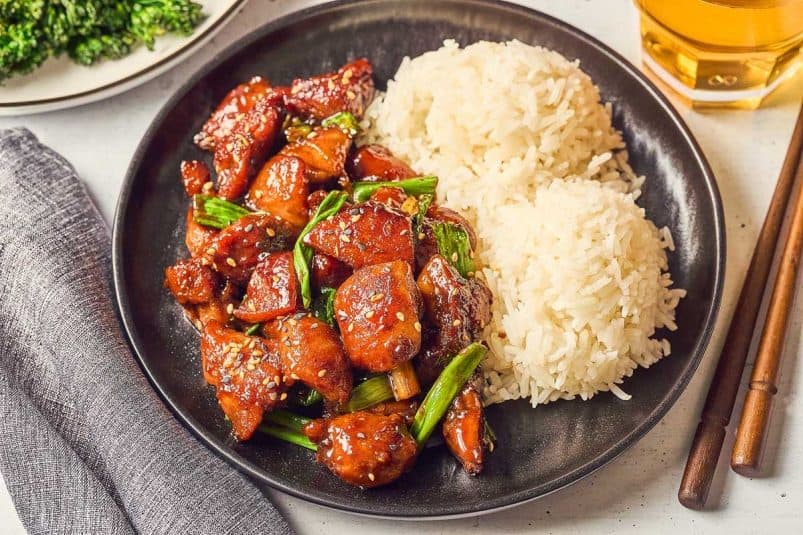 A plate featuring Five Spice Chicken Stir Fry with glazed chicken, green onions, and sesame seeds sits invitingly beside two servings of white rice. Chopsticks are elegantly placed alongside a glass of amber liquid, completing the scene.