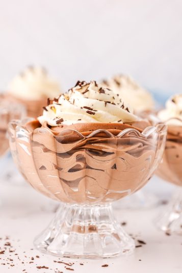 A close up picture of a glass dessert cup filled with Nutella chocolate cheesecake mousse with fresh whipped cream and chocolate shavings sprinkled about on a white counter top.