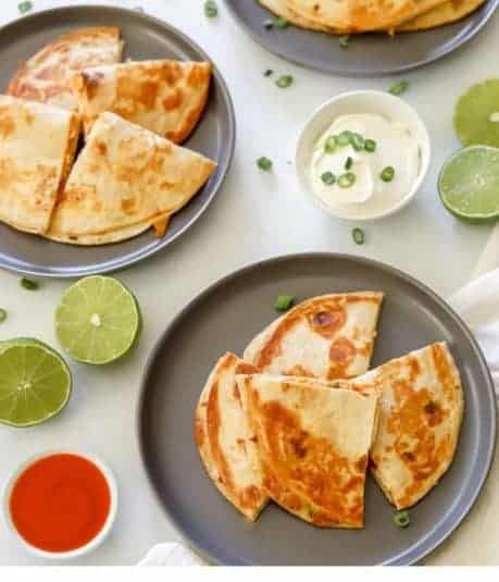 Two quesadillas cut into fours with a side of sour cream, buffalo wing sauce, and limes.