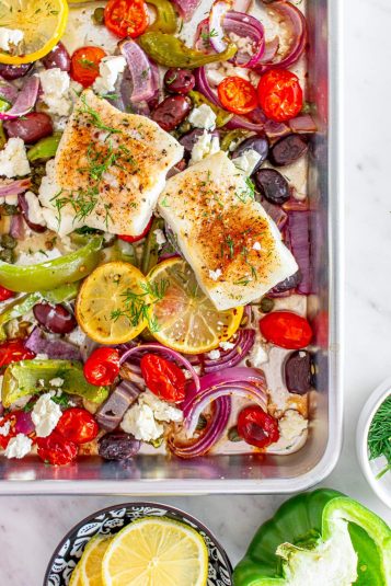 A baked Mediterranean cod with roasted vegetables, including tomatoes, bell peppers, and onions and garnished with lemon slices and fresh herbs.