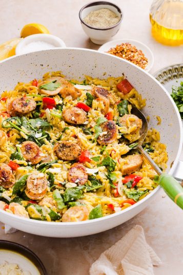 Photo of chicken sausage orzo styles in a large white pan, with a silver serving spoon with a green handle.