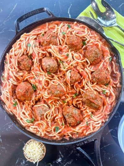 Spaghetti and Meatballs in a cast iron skillet garnished with fresh parsley and a small bowl of parmesan cheese for garnishing.