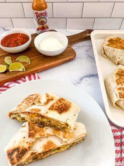 Homemade crunch wrap supremes on a white plate with a serving board with bowls of sour cream, salsa, a bottle of Tapatio, and lime slices in the background.