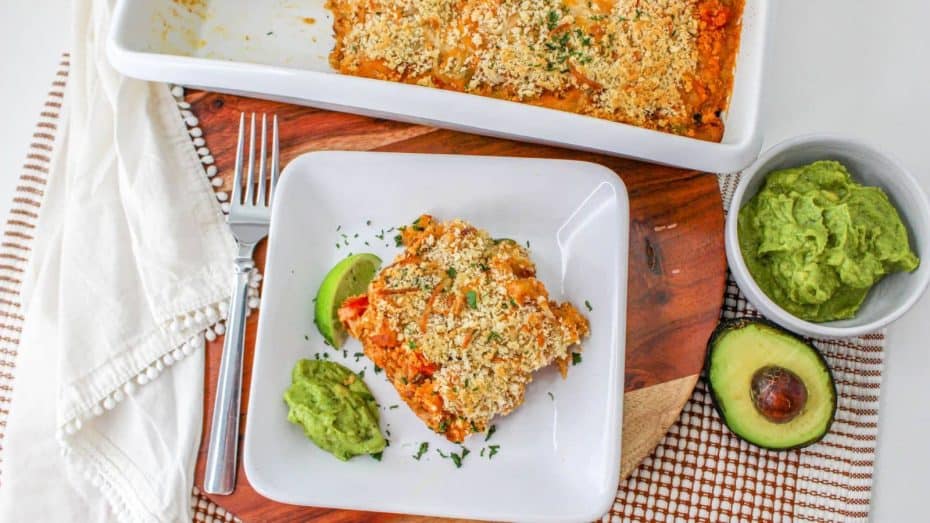 A serving of baked casserole with a crumbly topping on a plate, accompanied by guacamole and avocado slices. A casserole dish and more guacamole are in the background, placed on a wooden board.
