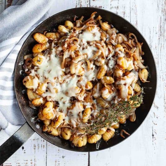 A cast iron skillet filled with French onion gnocchi with a bunch of thyme on the side.