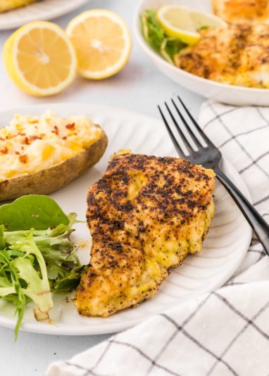 Lemon pepper fish served on white dish garnished with lemons and served with salad.