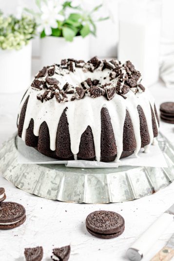A dark chocolate bundt cake with white icing drips and crushed oreos around the top of it.