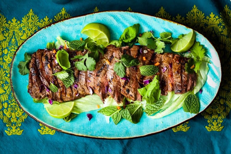 Flank steak salad with green herbs and lime wedges on a turquoise oval platter on patterned blue and green tablecloth.