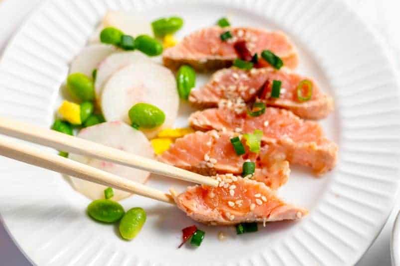 5 thin slices of salmon tataki on a white plate with scattered edamame and white radish on the left and a pair of chopsticks picking up a piece.