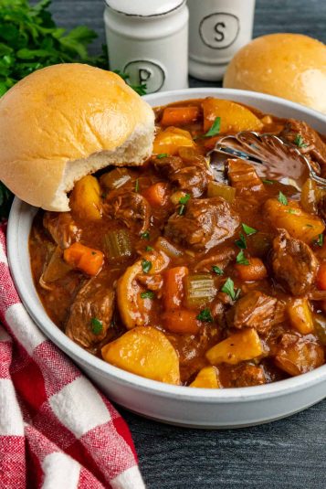 Slow cooker beef stew in a bowl served with a roll on the edge of the bowl and in the background.