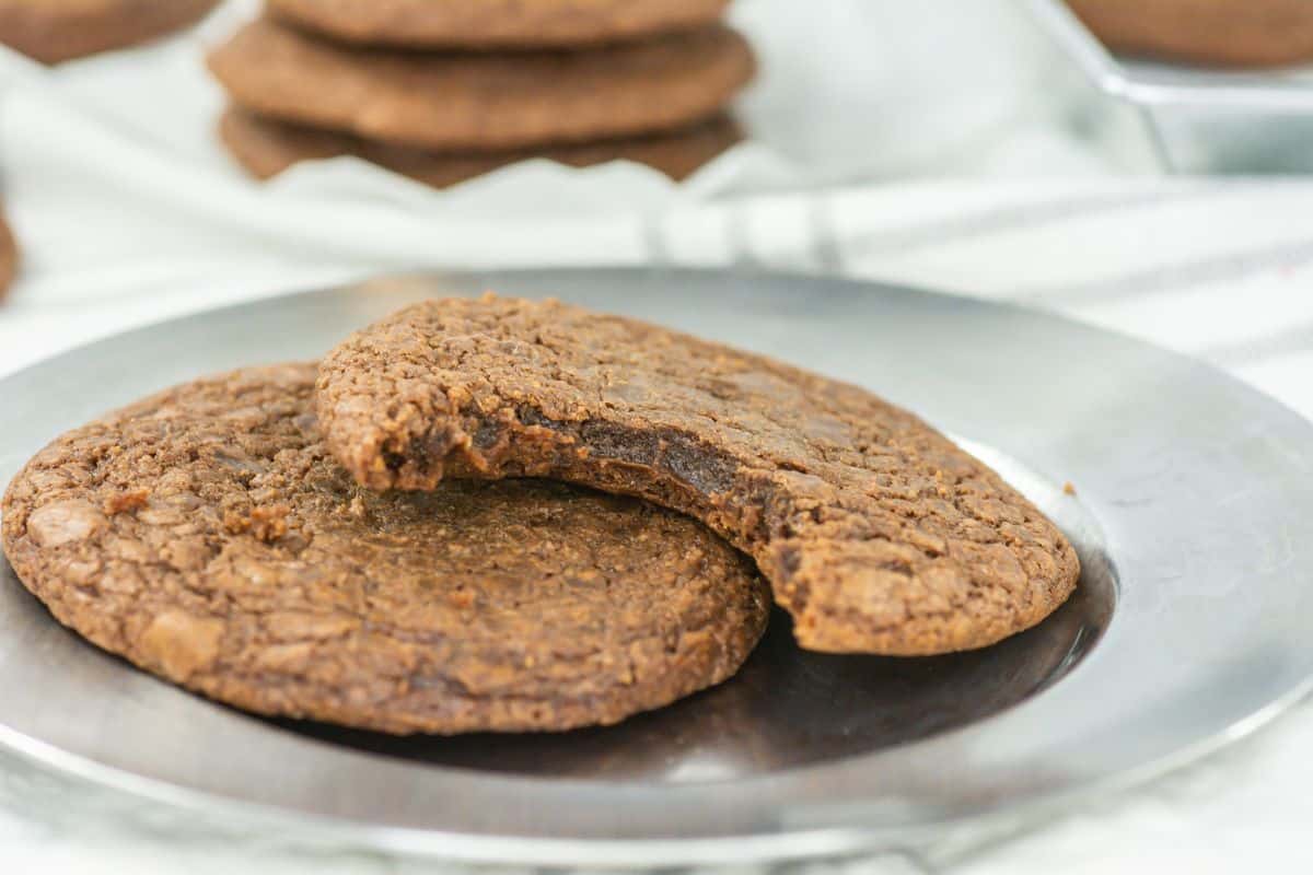 Two chocolate Nutella cookies are placed on a metal plate, with the cookie on top partially eaten. In the background, a blurred stack showcases more of these delicious treats made from an easy 3 Ingredient Nutella cookies recipe.