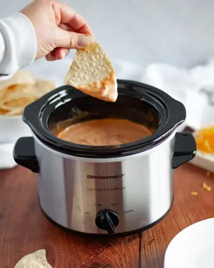 A hand holding a tortilla chip that was just dunked into the crockpot of hot chili cheese dip. additional tortilla chips, shredded cheese, plates, and a white linen sit around the crockpot.