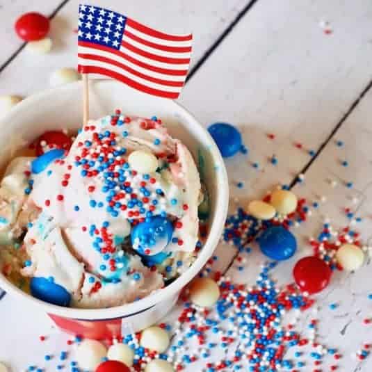 4th of July red white and blue ice cream in a flag cup with an American Flag pick, red, white and blue m & ms laying around with sprinkles.