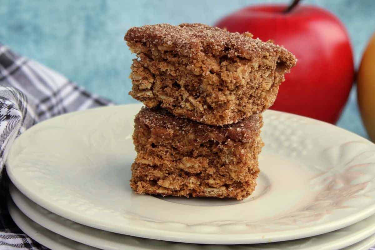 Two stacked square apple cider blondies on a white plate, with an out-of-focus red apple and folded plaid cloth in the background.