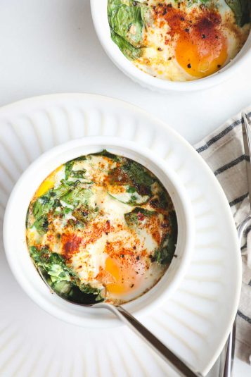 Two white ramekins with Baked Spinach and Egg Breafkast Cups with bright yellow yolks, green spinach and a pinch of red paprika.