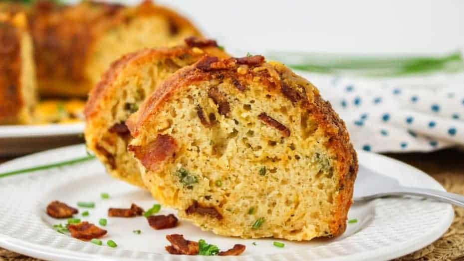 Two slices of bundt cake on a white plate, featuring visible bacon bits and green herbs, with a backdrop of blurred bread and a dotted napkin.