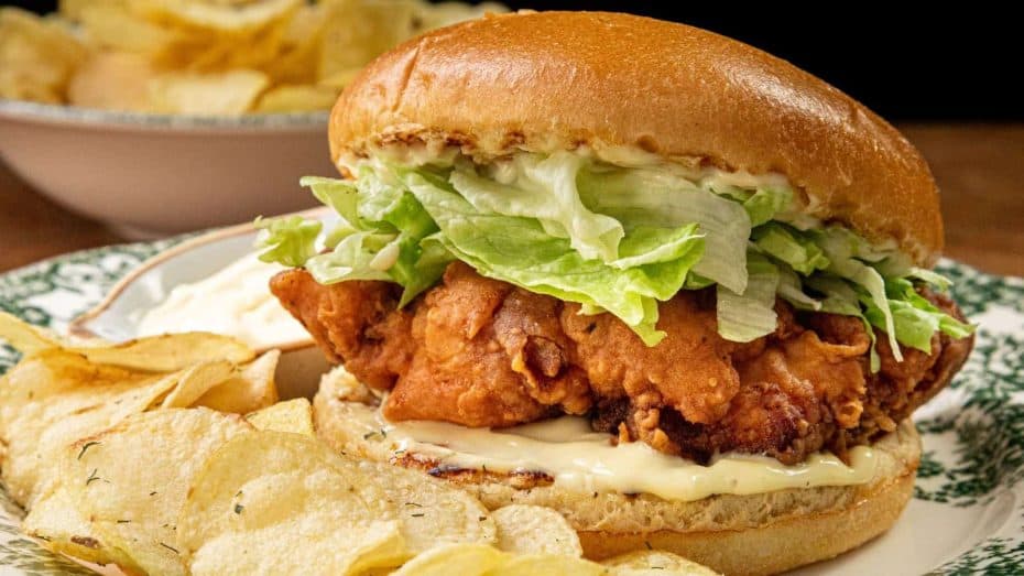 A fried chicken sandwich with lettuce and mayonnaise on a bun, served on a plate with potato chips.