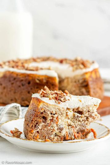 A single slice of Carrot Cake Cheesecake on a dessert plate with a view of the rest of the Carrot Cake Cheesecake and a glass bottle of milk in the background. Behind that is a white marble background.