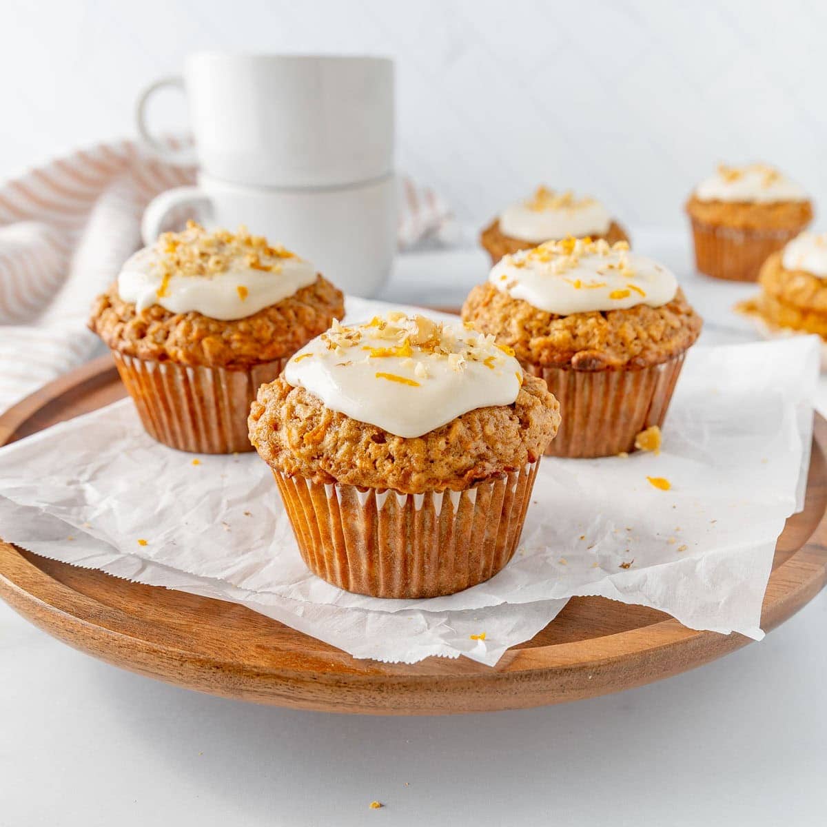 Muffins with white icing and orange zest on a wooden plate with white paper, two stacked white mugs in the background.