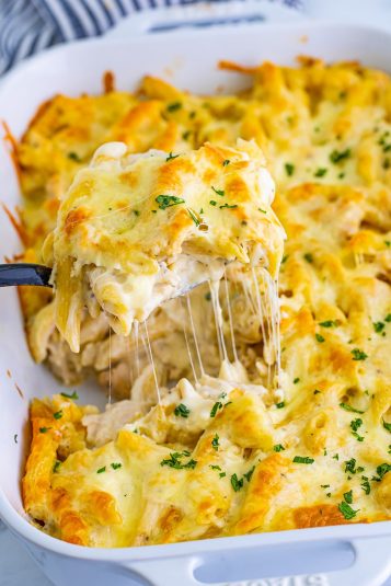 Chicken Alfredo Bake shown in a white casserole dish. A spoon is lifting up a portion with mozzarella cheese pull, navy striped linen in the background.