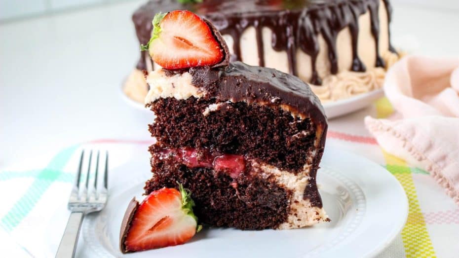 A slice of chocolate cake with a strawberry filling and dark chocolate glaze on a plate. Whole cake in the background, garnished with a halved strawberry. Fork on the side.