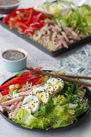 Cold noodle salad being lifted up with chopsticks, over a black round plate, with a black serving tray of more salad in background.
