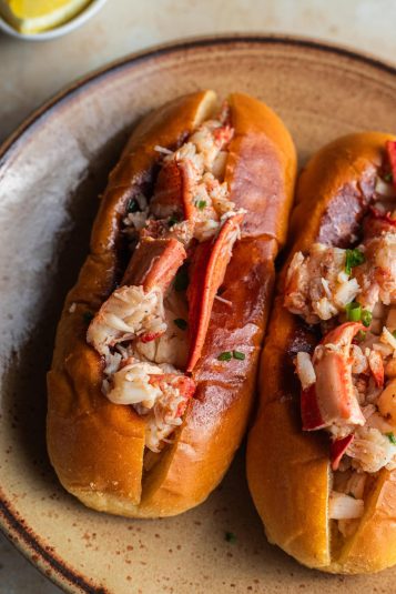 Two hot dog buns on a plate stuffed with lobster meat and garnished with chives.