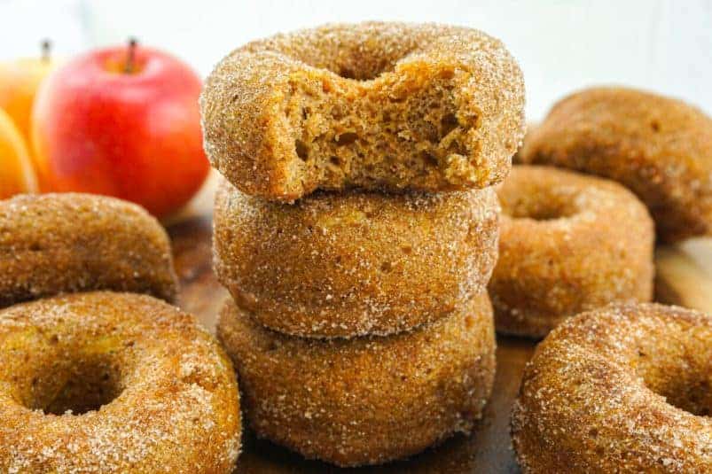 Three apple cider breakfast doughnuts stacked on top of each other with a bite out of the top doughnut. More doughnuts and apples in the background.