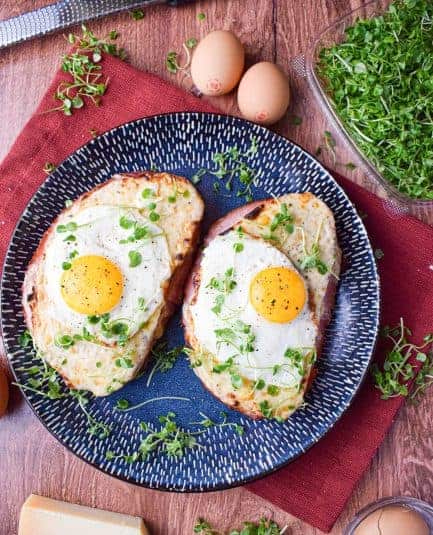 Overhead shot of 2 croque madame sandwiches topped with gruyere cheese, a fried egg and greens.