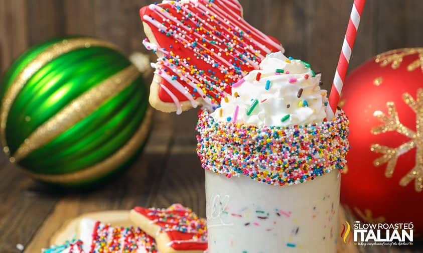 A milkshake topped with whipped cream and colorful sprinkles, a sugar cookie, and a red and white striped straw.