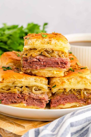 A plate filled with small french dip sandwiches. One sandwich is on top of the others.