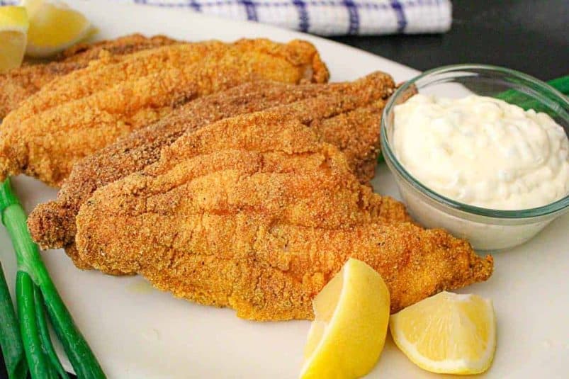 Fried catfish filets with tartar sauce in a ramekin and lemons on the side.