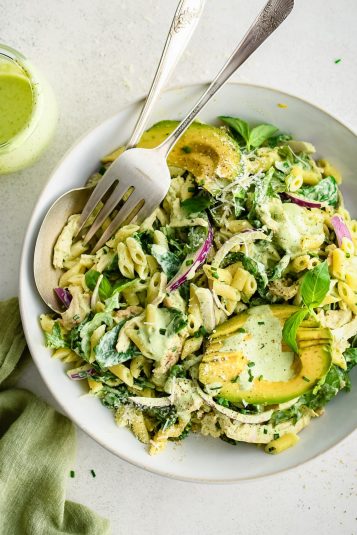 Large white serving bowl filled with creamy green goddess pasta salad made with short penne pasta, shredded chicken, sliced red onion, fresh spinach, arugula, and avocado all tossed in homemade green goddess dressing.