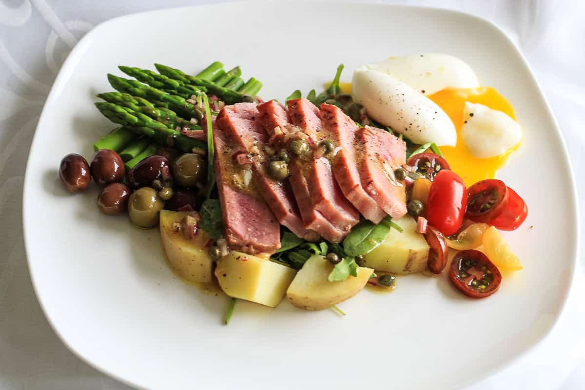 Nicoise salad composed of rare sliced tuna, nicoise olives, and runny egg arranged on a white plate.