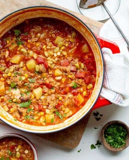 Red-handled Dutch oven with Shortcut Brunswick Stew on a wooden board with a white kitchen towel, a bowl of stew, and a small wooden bowl with chopped parsley.