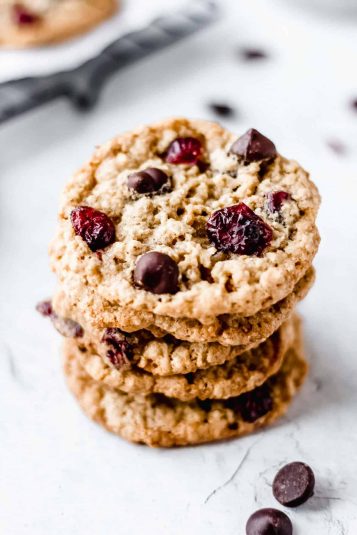 Stack of 5 oatmeal cranberry chocolate chip cookies on a white background with part of a cooling rack and extra chips and cranberries in the background.