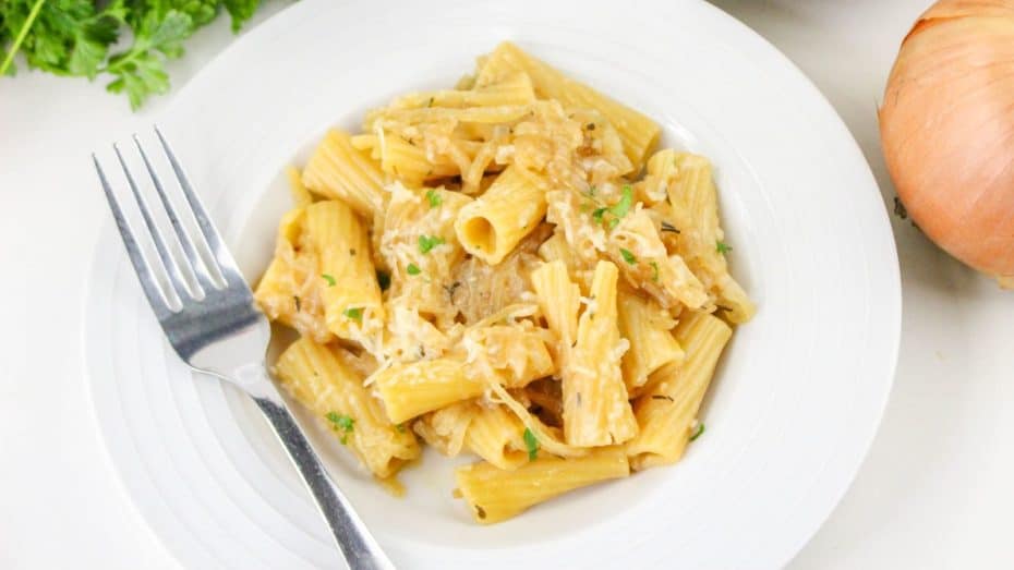 A plate of rigatoni pasta with shredded cheese and herbs, next to a fork on a white dish. An onion and parsley are in the background.