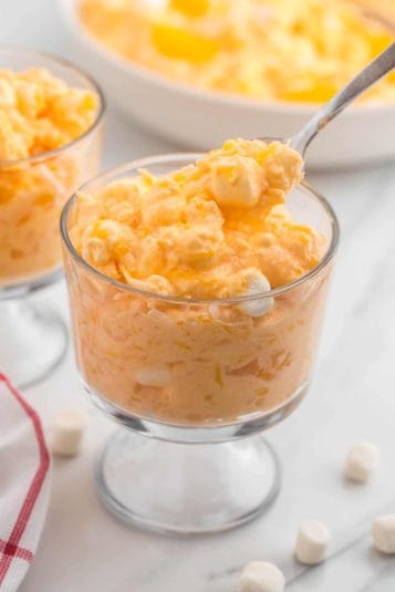 A footed dessert dish filled with a serving of orange fluff jello salad. It's being enjoyed with a spoon.