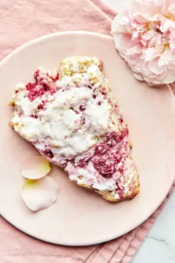 A raspberry scone on a small pink plate with a pale pink rose and two pink rose petals nearby.