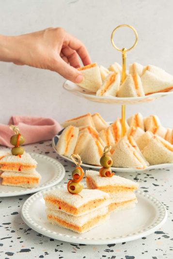 Two servings of Sandwichitos de Mezcla are on a white plate with a bead rim in the foreground. A hand reaches for a sandwich of a serving tray in the background.