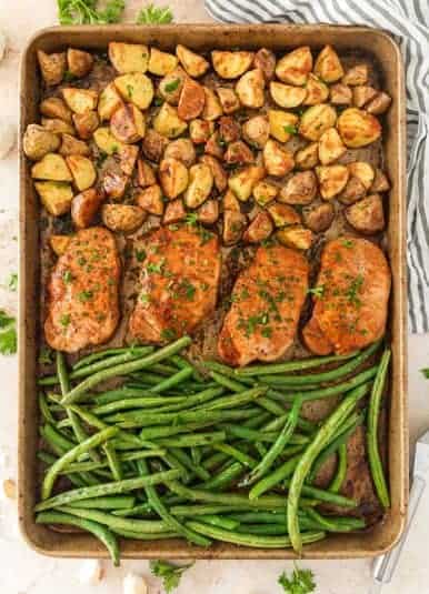 Sheet pan with baked pork chops, potatoes and green beans topped with chopped fresh parsley.
