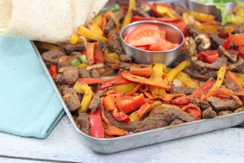 Baking tray with cooked steak, bell peppers, and veggies with a steel bowl of fresh tomatoes in the middle and flour tortillas folded to the side.