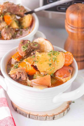 White bowl filled with slow cooker beef stew containing chunks of beef, potatoes, carrots, and garnished with fresh herbs. A wooden pepper mill is in the background.
