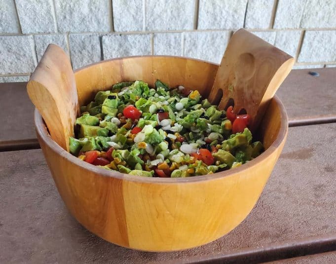 Image shows a wooden bowl containing avocado tomato salad shown from overhead and sitting on a picnnic table.