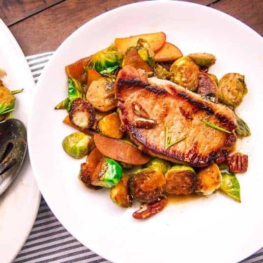 Pork chop on a bed of brussels sprouts and apples on a white plate.