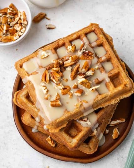 A stack of carrot cake waffles topped with cream cheese maple syrup and chopped pecans.